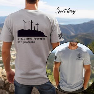 Peut inclure: T-shirt gris sport avec un graphisme noir de trois croix sur une colline et le texte "y'all need Proverbs not pronouns." Un drapeau américain est sur la manche. Le t-shirt présente également un logo de lion.