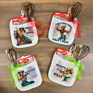 May include: Four white oven mitts with cowboy-themed graphics, each holding a Betty Crocker cookie mix packet, a wooden spoon, and a whisk. The mitts are tied with red and green ribbons. Text on the mitts includes phrases like "Horses Keep Me Stable" and "I do a thing called what I want."
