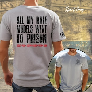 May include: Sport grey t-shirt with the text "ALL MY ROLE MODELS WENT TO PRISON" in black, and names in red. An American flag is on the sleeve. A smaller image shows a similar shirt with a lion logo.