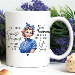 May include: White ceramic mug with a black and white illustration of a woman in a blue polka dot dress and a blue bow in her hair. The text on the mug reads "The term 'Domestic Housewife' implies that there are Feral Housewives and now I have a new life goal."