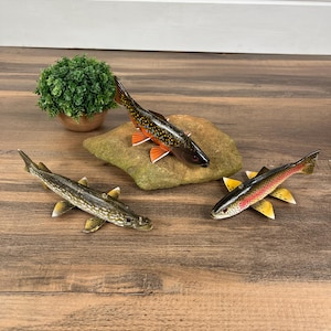 May include: Three wooden fish sculptures in various colors and patterns are displayed on a wooden surface. One fish is dark with orange fins, another is green and white, and the third is red, black, and yellow.