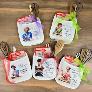 Może przedstawiać: Białe rękawice kuchenne z ilustracjami i tekstem w stylu retro, każda z trzepaczką, szpatułką i mieszanką do pieczenia Betty Crocker. Projekty obejmują kobietę piekącą i frazy takie jak "Pieczenie, ponieważ morderstwo jest złe."