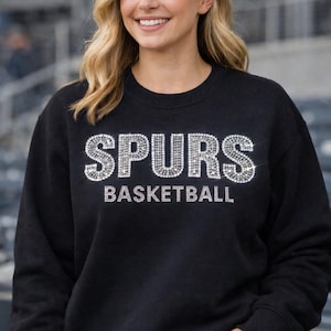 May include: Black crewneck sweatshirt with the word "SPURS" and the word "BASKETBALL" in silver rhinestones. The sweatshirt is worn by a person with blonde hair.