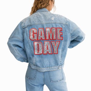 May include: Light blue denim jacket with the words "GAME DAY" in silver sequins outlined in red on the back. The jacket has silver embellishments on the shoulders and is paired with matching jeans.