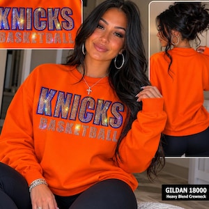 May include: An orange crewneck sweatshirt with the text "KNICKS BASKETBALL" in sparkling rhinestones. The sweatshirt has a ribbed collar and cuffs. The image also shows a close-up of the text and the back of the sweatshirt.