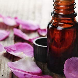 May include: A brown glass bottle with a white rim and a black cap sits on a wooden surface, surrounded by pink and white rose petals. The bottle is likely for essential oils or perfumes.