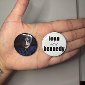 May include: Two circular badges. One displays a portrait of a person with light-colored hair and a blue jacket. The other is white with the text "leon slut kennedy" in black and blue.