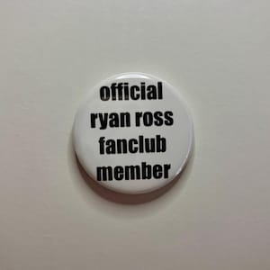 May include: A white button badge with the text "official ryan ross fanclub member" in black. The badge is circular and has a shiny surface. The text is in a bold, sans-serif font and is centrally aligned.
