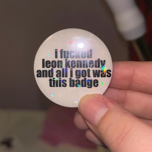 May include: A circular, cream-coloured badge featuring the black text "i fucked leon kennedy and all i got was this badge". The badge has a holographic, glittery finish with small, colourful star accents.