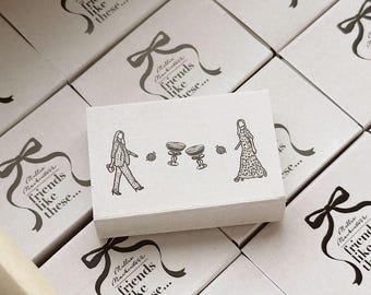 Pode incluir: Uma caixa retangular de papelão branco com um desenho de linha preta de duas figuras, duas taças e duas pequenas esferas. O texto Millie Mackintosh's friends like these... está impresso em um gráfico de fita preta na superfície da caixa.