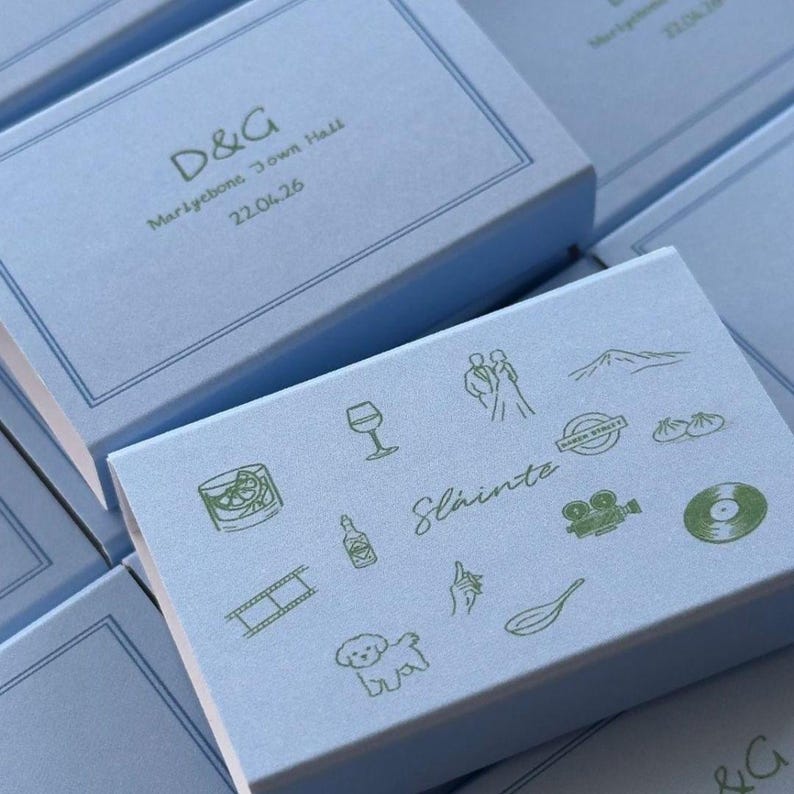 May include: A light blue rectangular box featuring dark green line art icons including a wine glass, a couple, a mountain, a film strip, a bottle, the word Sl&aacute;inte, a camera, a vinyl record, a dog, a hand, and a whisk, with a double-line border.