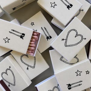 May include: A collection of rectangular matchboxes with a cream-colored exterior. Each box features black line art illustrations of either a heart pierced by an arrow or two parallel arrows accompanied by stars. One open box reveals pink-tipped matches.