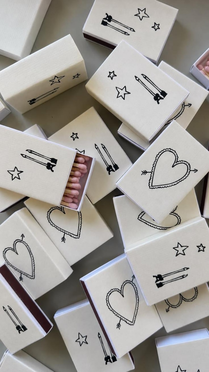 May include: A collection of rectangular matchboxes with a cream-colored exterior. Each box features black line art illustrations of either a heart pierced by an arrow or two parallel arrows accompanied by stars. One open box reveals pink-tipped matches.