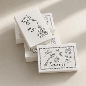 May include: A stack of white rectangular matchboxes featuring black line art illustrations. One box shows a cherub pouring liquid into glasses with the text CIN CIN. Another box displays a collage of a disco ball, champagne, and the date 07.03.25.