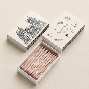 May include: Two rectangular matchboxes with off-white covers and brown strike strips. One cover features a black line drawing of a building, the other shows icons of a cake, ring, bird, bottle, and the text "Majlaa & Thomas 15.06.2026". Matches have light wood stems and brown tips.