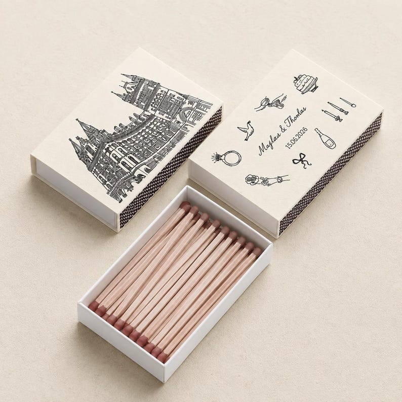 May include: Two rectangular matchboxes with off-white covers and brown strike strips. One cover features a black line drawing of a building, the other shows icons of a cake, ring, bird, bottle, and the text "Majlaa & Thomas 15.06.2026". Matches have light wood stems and brown tips.