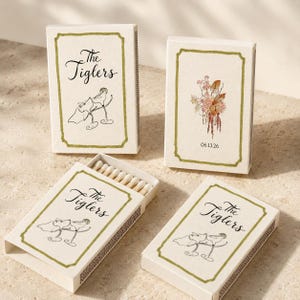 May include: Four rectangular matchboxes with off-white covers featuring a green border. Three boxes display the text The Tiglers above a line drawing of two champagne glasses. One box shows a floral illustration and the date 06.13.26.