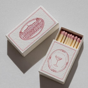 May include: Two rectangular matchboxes with a textured white surface and red ink illustrations. One box is closed, showing a building in an oval frame. The other is open, revealing wooden matches with pink tips and a martini glass illustration.