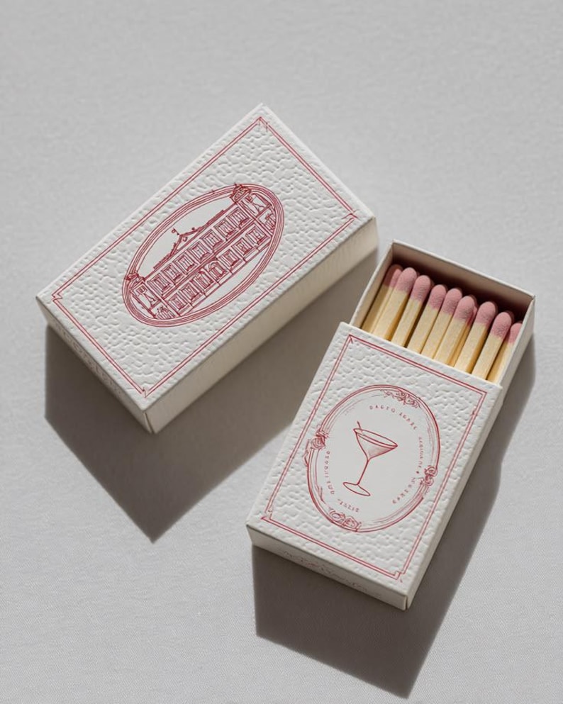 May include: Two rectangular matchboxes with a textured white surface and red ink illustrations. One box is closed, showing a building in an oval frame. The other is open, revealing wooden matches with pink tips and a martini glass illustration.