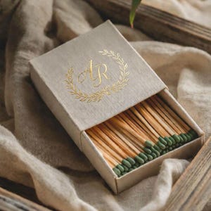 Puede incluir: Una caja de cerillas beige con un monograma dorado y un diseño de corona de laurel. La caja abierta revela cerillas de madera con puntas verdes. Las cerillas están cuidadosamente dispuestas dentro de la caja.
