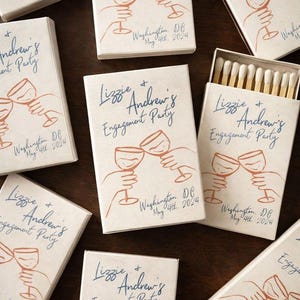 May include: Off-white rectangular matchboxes featuring blue cursive text reading Lizzie + Andrew's Engagement Party and Washington, DC May 4th, 2024. A line art illustration in orange depicts two hands holding wine glasses in a toast.