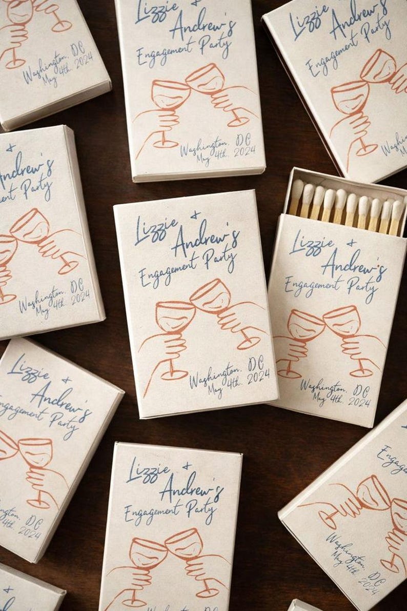 May include: Off-white rectangular matchboxes featuring blue cursive text reading Lizzie + Andrew's Engagement Party and Washington, DC May 4th, 2024. A line art illustration in orange depicts two hands holding wine glasses in a toast.