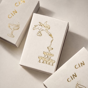 May include: Three rectangular white boxes with gold foil stamping. The central box features a winged cherub pouring liquid into a stacked pyramid of champagne coupes. The other two boxes display the text CIN CIN and a single champagne coupe illustration.