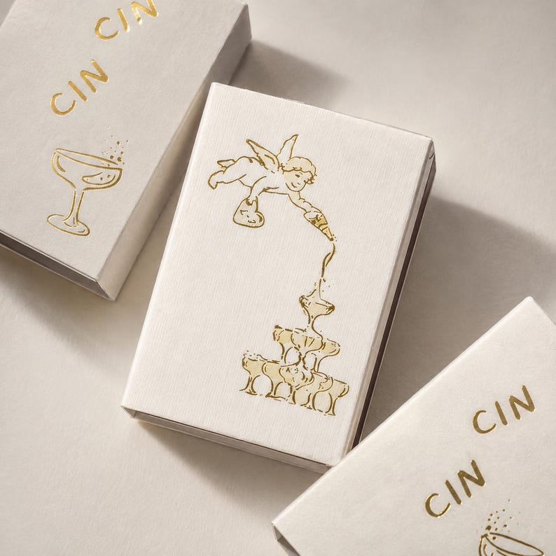 May include: Three rectangular white boxes with gold foil stamping. The central box features a winged cherub pouring liquid into a stacked pyramid of champagne coupes. The other two boxes display the text CIN CIN and a single champagne coupe illustration.