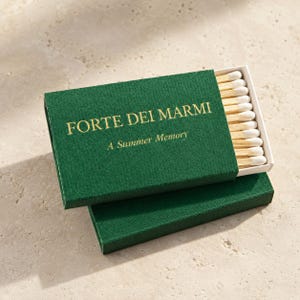 May include: A rectangular green matchbox with a sliding tray containing wooden matches with white tips. The top of the box features gold text that reads FORTE DEI MARMI and A Summer Memory. The box is resting on top of a matching green box base.