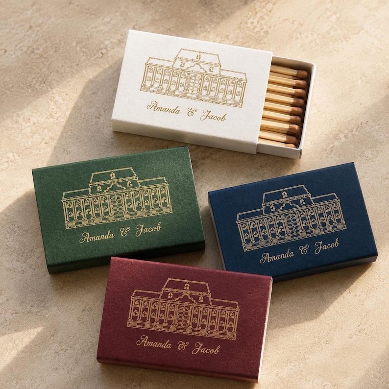 May include: Four rectangular matchboxes in white, dark green, navy blue, and burgundy. Each box features a gold foil line drawing of a building and the text Amanda & Jacob. The white box is partially open, revealing wooden matches inside.