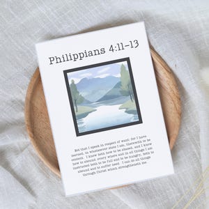 May include: A white card with the text "Philippians 4:11-13" and a landscape painting in a black frame. The card also includes a Bible verse. The card is on a wooden plate.