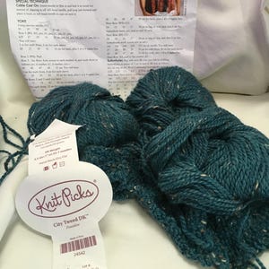 May include: Two skeins of teal blue tweed yarn with small flecks of white and brown. A white paper label attached to the yarn reads Knit Picks City Tweed DK Poseidon, 55% Merino Wool, 25% Superfine Alpaca, 20% Donegal Tweed. A knitting pattern sheet is included.