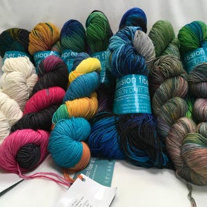 May include: A collection of colorful yarn skeins in various hues, including blue, green, yellow, pink, and black. Some skeins have labels that read "Expression Fiber Arts". The yarn is arranged on a white surface.
