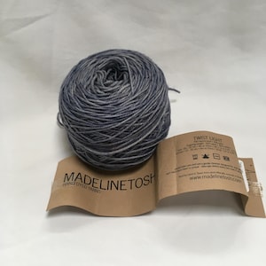 Può includere: Un gomitolo di filato Twist Light blu e grigio, con un'etichetta di carta marrone che recita "MADELINETOSH HAND DYED YARNS". L'etichetta include anche il testo "TWIST LIGHT".