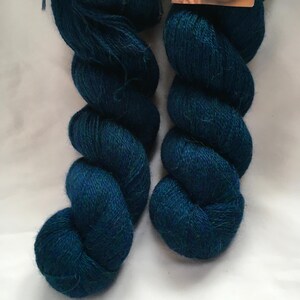 Puede incluir: Dos madejas de hilo Alpaca Lace de color verde azulado se muestran sobre un fondo blanco. El hilo est&aacute; enrollado firmemente, mostrando su textura y color. Las etiquetas en la parte superior dicen "Alpaca Lace".