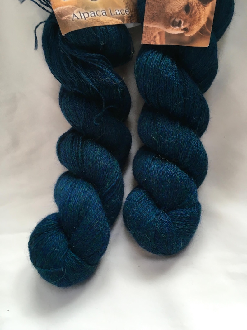 Puede incluir: Dos madejas de hilo Alpaca Lace de color verde azulado se muestran sobre un fondo blanco. El hilo est&aacute; enrollado firmemente, mostrando su textura y color. Las etiquetas en la parte superior dicen "Alpaca Lace".