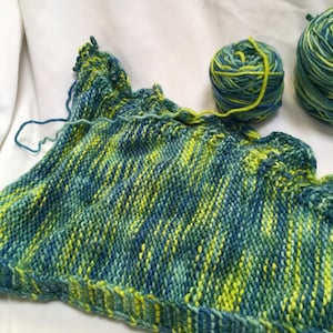 May include: A knitted item in progress, featuring blue and yellow variegated yarn, with a decorative ruffled edge. Two balls of yarn in complementary colours are also present. The project is displayed on a white background.