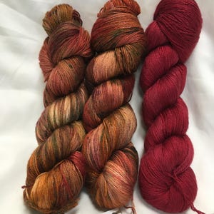 May include: Three skeins of yarn in shades of orange, brown, and red. Two skeins have a variegated color pattern, while the third is a solid deep red. The yarn is displayed on a white surface. The labels read "Autumn Lake" and "Red Velvet Rose".