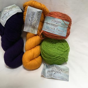 Könnte beinhalten: Eine Sammlung bunter Garnknäuel in tiefem Lila, Goldgelb, Orange und Grün. Jedes Knäuel trägt ein Etikett mit der Aufschrift "Heritage Silk Paints" und "Cascade Yarns". Das Garn besteht aus Merino Superwash Wolle und Maulbeerseide.
