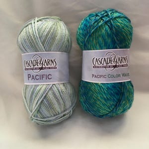 Op de afbeelding: Twee strengen Cascade Yarns Pacific fijn garen. Eén is een lichtblauwe en groene mix, met het label "Pacific". De andere is een turquoise, blauwe en gele mix, met het label "Pacific Color Wave".