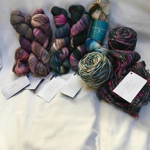 May include: A collection of colorful yarn skeins and balls in various shades of pink, green, purple, and cream. Several skeins are arranged together, with tags attached. A teal paper tube with text is also visible, along with a small knitted piece.