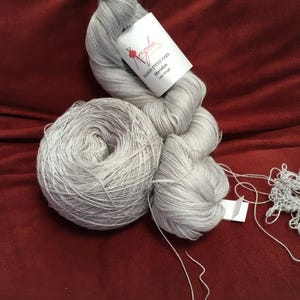 May include: Three skeins of gray hand-dyed yarn, one in a ball, one in a loose bundle, and one partially unraveled. A white label reads "Anzula Hand Dyed Yarn Meridian Lace Weight". The yarn is on a red surface.
