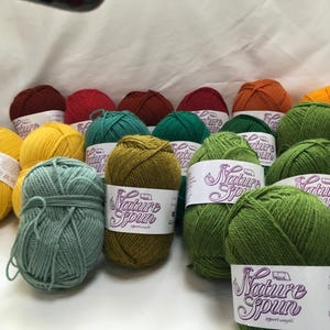 Può includere: Una collezione di gomitoli di lana colorati in varie tonalità, tra cui giallo, verde, arancione e rosso. Ogni gomitolo è etichettato con "Nature Spun" ed è adatto per progetti di maglieria o uncinetto. La lana è esposta su una superficie bianca.