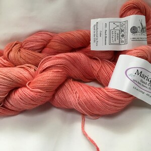 May include: Peach-colored yarn skeins with labels. The labels indicate the yarn is hand-dyed, made of baby alpaca, and the color is Peach Blossoms. The yarn is 3.5 oz (100g) and 400 yards. The yarn is manufactured in Peru and dyed in the USA.