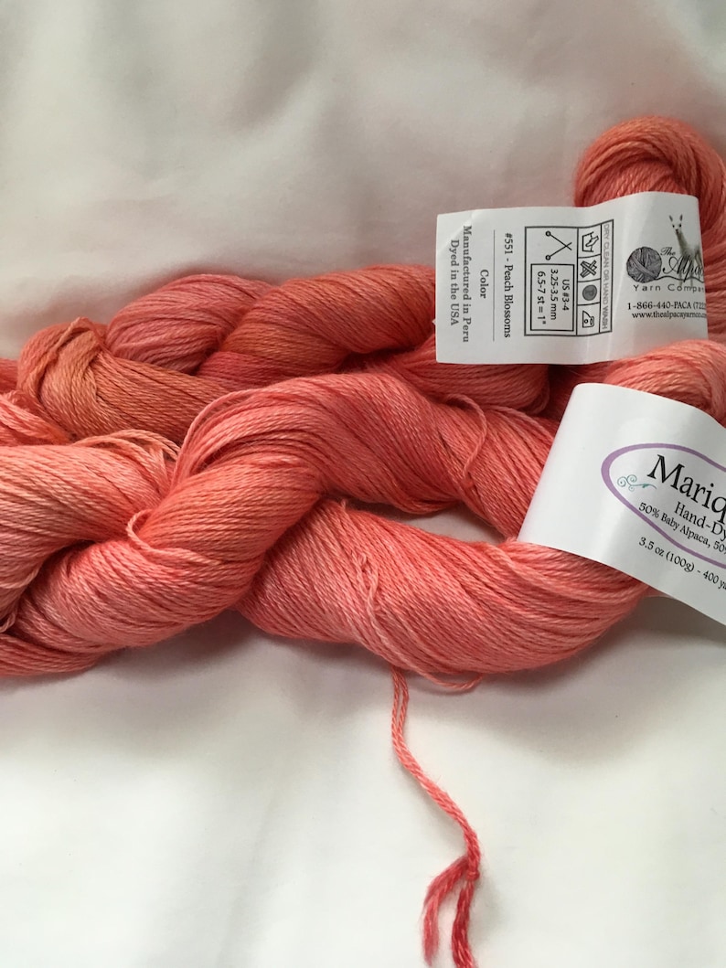 May include: Peach-colored yarn skeins with labels. The labels indicate the yarn is hand-dyed, made of baby alpaca, and the color is Peach Blossoms. The yarn is 3.5 oz (100g) and 400 yards. The yarn is manufactured in Peru and dyed in the USA.
