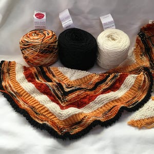 May include: Three balls of yarn in orange, black, and white, alongside a knitted shawl in similar hues. The shawl features a striped design with a black scalloped border. The yarn is labelled "Yummy 2-Ply" by Miss Babs.