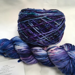May include: A skein of Unshaken Enduring Worsted yarn in shades of blue, purple, and black. The yarn is a blend of superwash merino wool and nylon. A sample of the yarn is displayed next to the skein. A tag with the yarn details is visible.