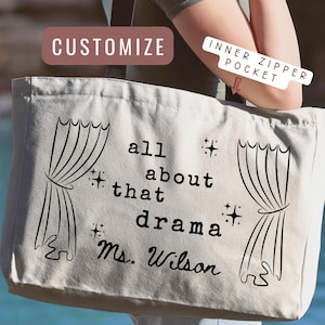 May include: A beige tote bag with black text that reads "all about that drama Ms. Wilson" and curtain graphics. The bag has an inner zipper pocket and is being held outdoors.
