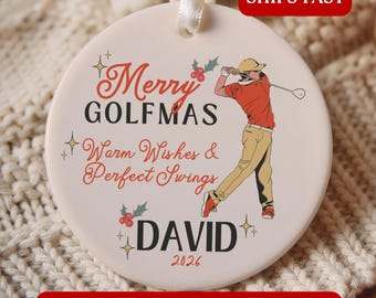 May include: A round white ceramic ornament featuring a graphic of a golfer in an orange shirt and tan trousers. The text reads Merry GOLFMAS, Warm Wishes & Perfect Swings, DAVID, and 2026. The design includes small holly berries and star accents.