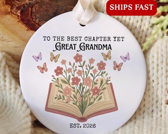 May include: A round white ceramic ornament with a cream ribbon, featuring an open book illustration with pink flowers emerging from the pages, surrounded by colourful butterflies, with the text TO THE BEST CHAPTER YET GREAT GRANDMA EST. 2026.